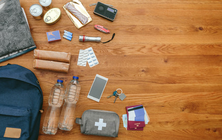 Das Bild zeigt einen Tisch, auf dem mehrere Gegenstände zu sehen sind: Wasserflaschen, Handy, Schlüssel, Pflastertasche, Tabletten, Streichhölzer, Taschenlampe, ein Seil und ein kleines Radio.